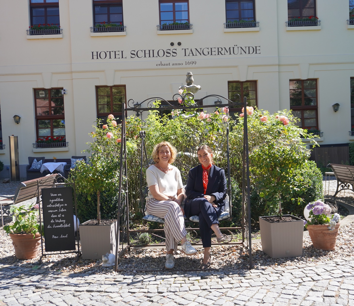 Eine freundlich lächelnde Dame sitzt auf einer schmiedeeisernen Hollywoodschaukel. zwischen Rosensträuchern. Neben ihr sitzt eine Firmenkundenberaterin der KSK Stendal. Im Hintergrund ist das Hotel Schloss Tangermünde zu sehen.
