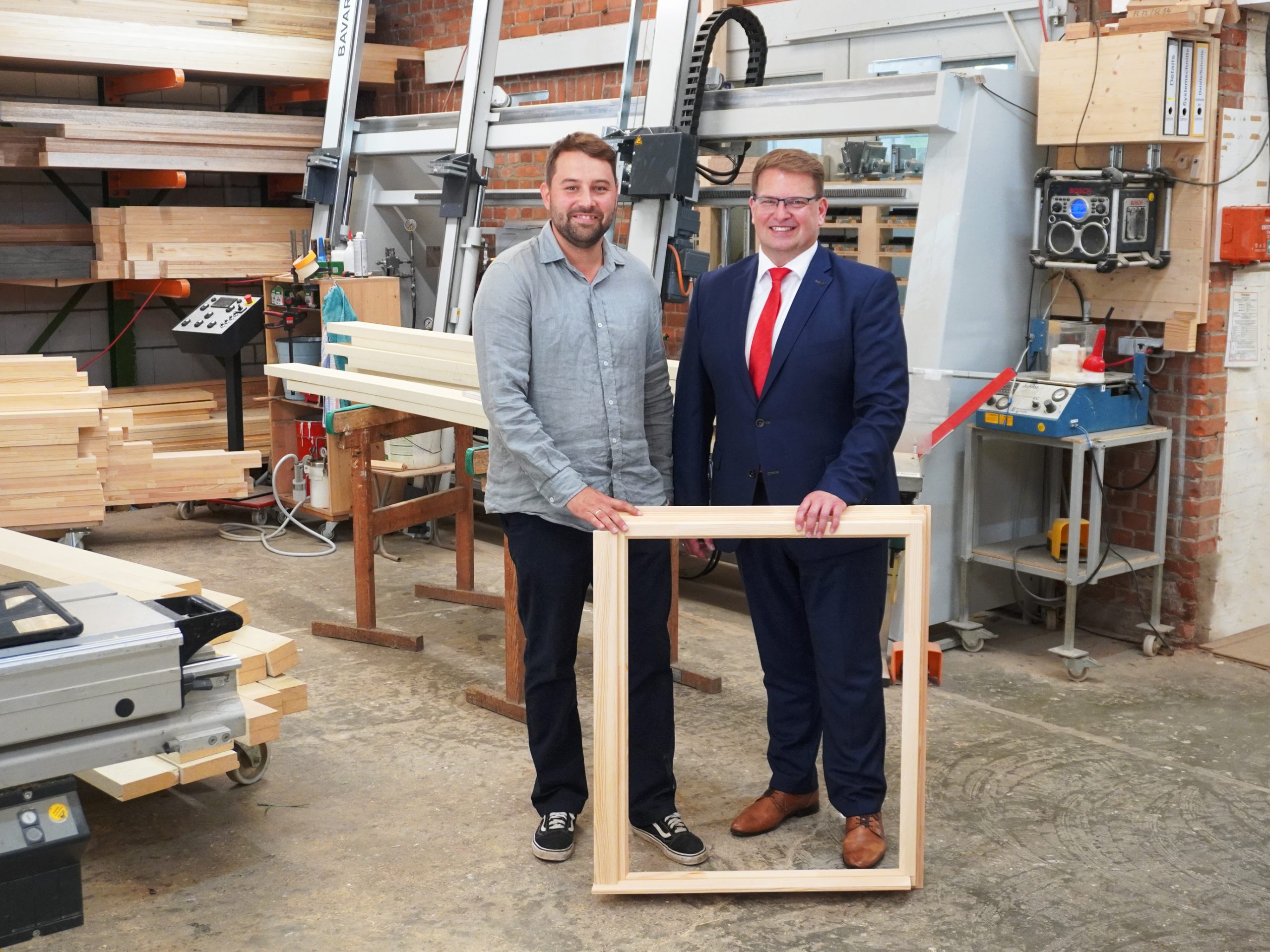 Ein freundlich lächelnder Herr steht in einer Werkstatt und legt seine Hand an einen Holzfensterrahmen. Neben ihm steht ein ebenfalls lächelnder Firmenkundenberater der KSK Stendal.